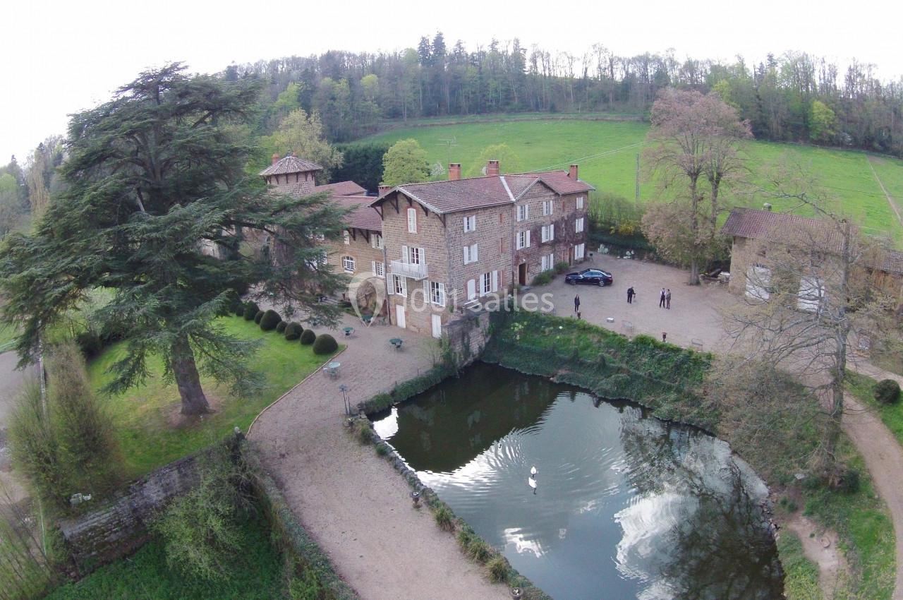 Vue aérienne d'une grande maison en pierre près d'un étang entouré de verdure et d'arbres.