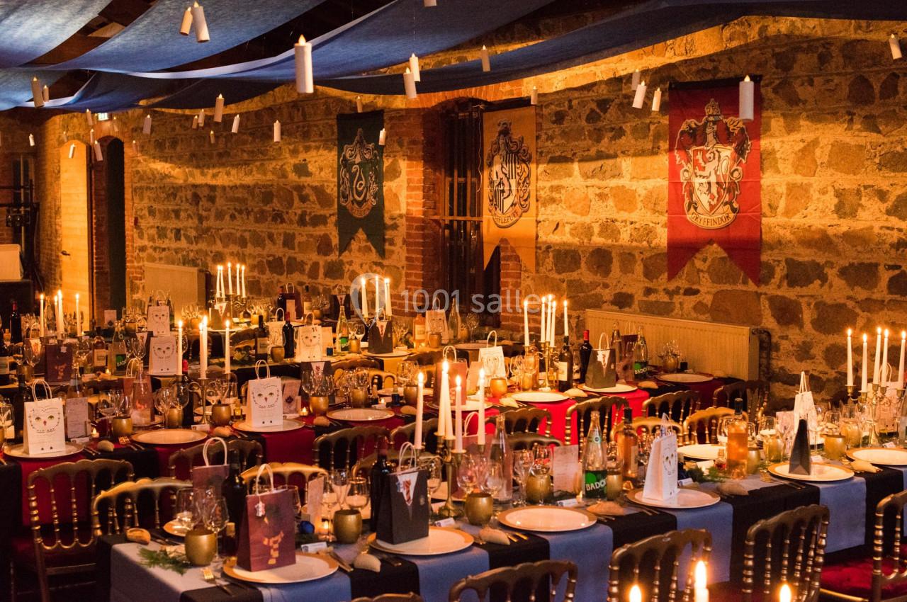 Salle décorée pour un banquet avec des tables dressées, des bougies suspendues et des bannières aux murs en pierre.