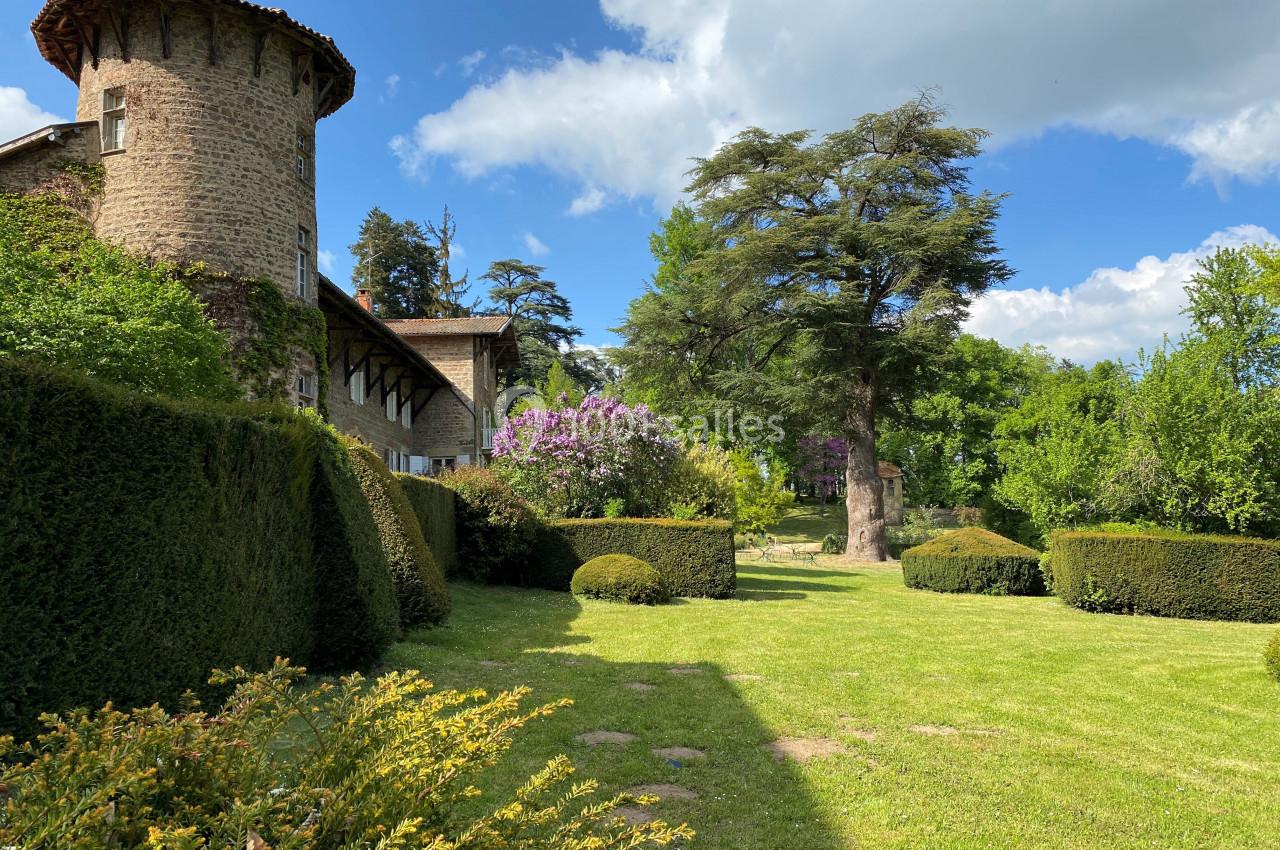 Jardin verdoyant avec pelouse, arbustes taillés, arbres imposants et bâtiment en pierre avec tour ronde sous un ciel bleu.