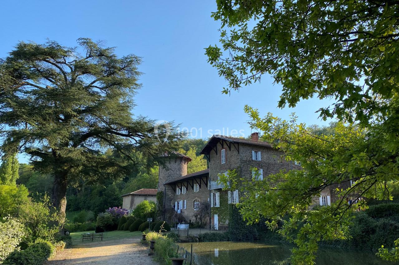 Grande maison en pierre entourée de verdure, avec un étang au premier plan et un grand arbre sur la gauche.