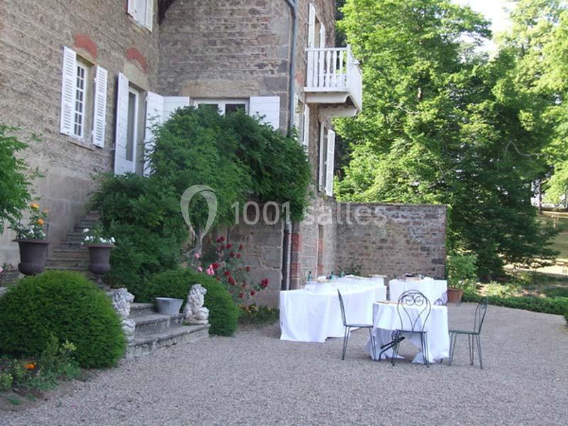 Terrasse d'une maison en pierre avec tables dressées, chaises en fer forgé et végétation environnante.