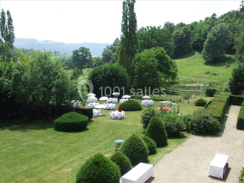 Vue d'un jardin verdoyant avec des tables rondes dressées sous des parasols blancs, entouré de collines et d'arbres.