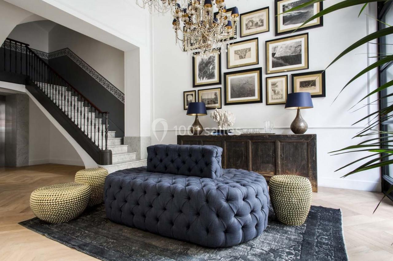 Salon élégant avec canapé capitonné gris, poufs tressés, tapis sombre, buffet en bois, cadres au mur et lustre imposant.