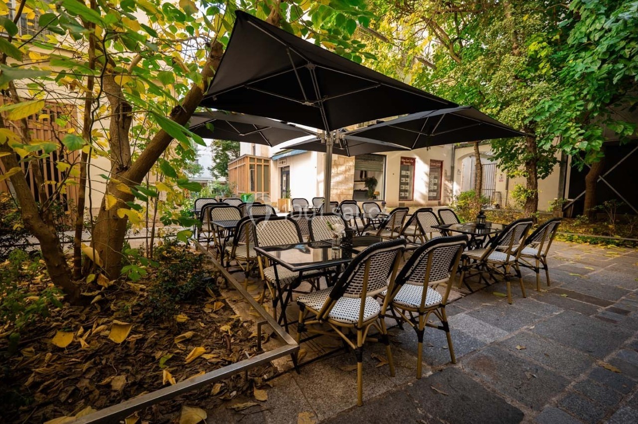Terrasse extérieure avec tables et chaises en osier sous de grands parasols noirs, entourée de végétation.