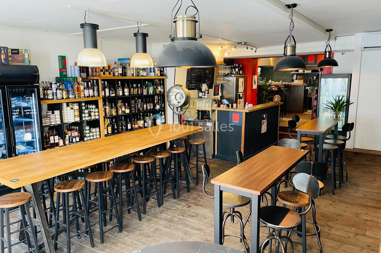 Salle de restaurant avec tables en bois, chaises hautes, étagères remplies de bouteilles et comptoir en arrière-plan.