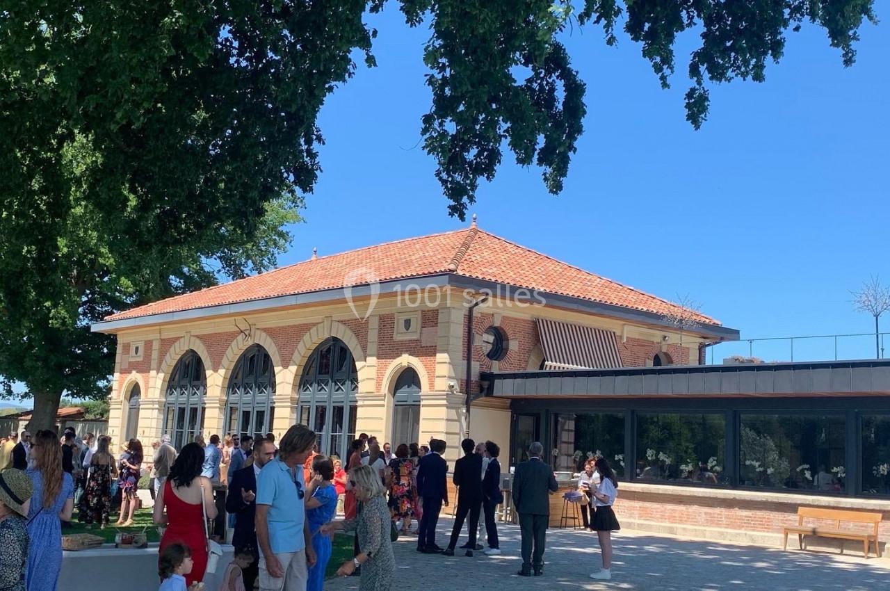 Location salle Cleppé (Loire) - L'Orangerie de Châtel #10 Groupe de personnes rassemblées devant un bâtiment en briques avec de grandes arches, sous un ciel bleu et des arbres.