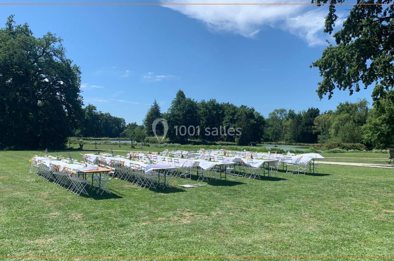 Location salle Cleppé (Loire) - L'Orangerie de Châtel #8 Tables et chaises blanches disposées en extérieur sur une pelouse entourée d'arbres sous un ciel partiellement dégagé.