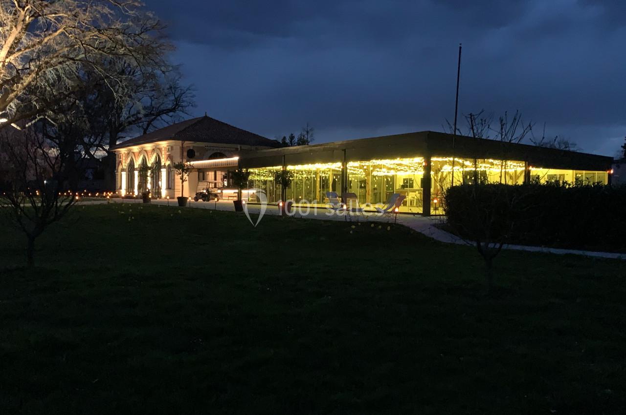 Location salle Cleppé (Loire) - L'Orangerie de Châtel #18 Bâtiment éclairé avec des guirlandes lumineuses, entouré d'arbres et d'une pelouse, sous un ciel sombre.