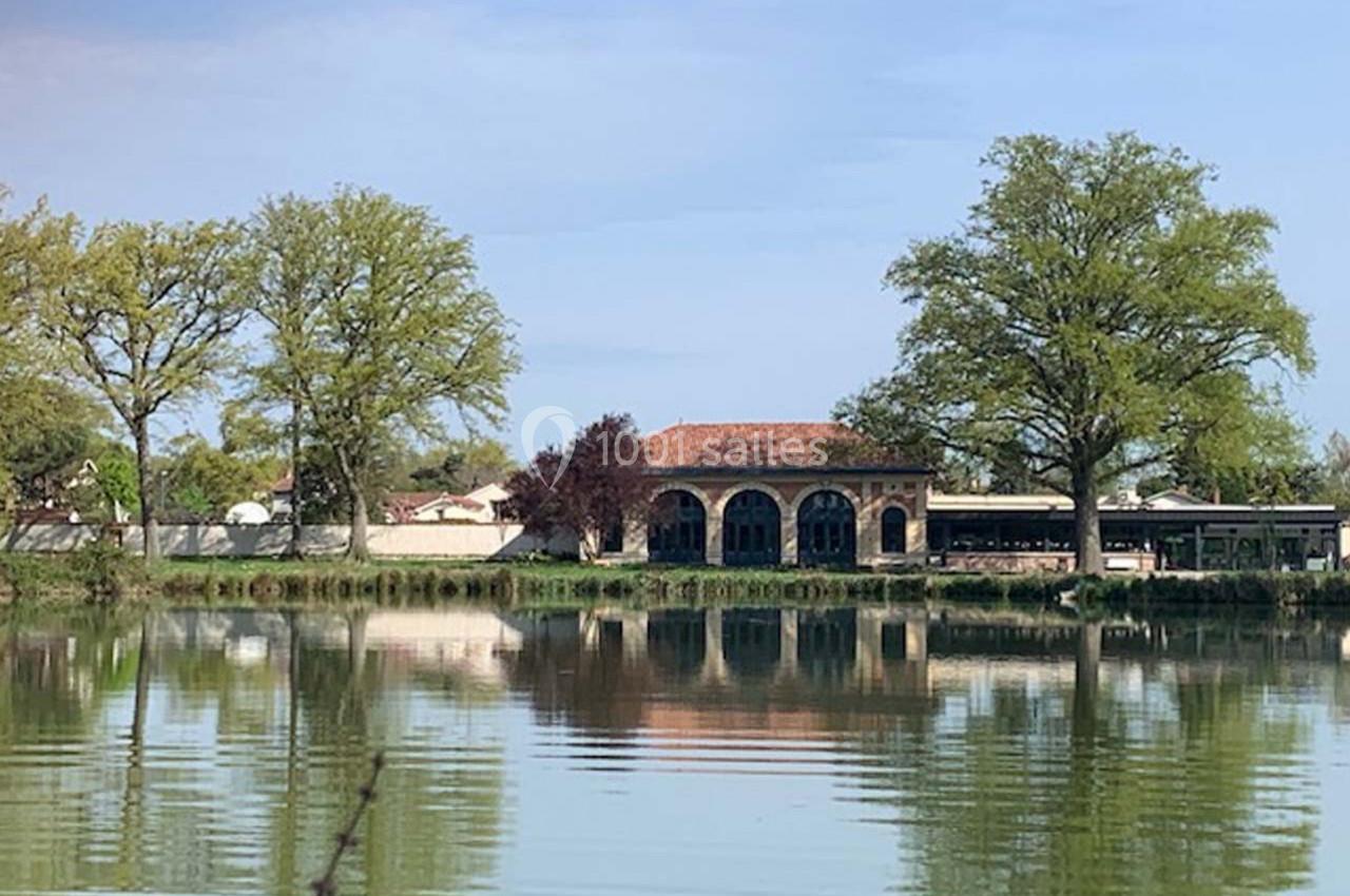Location salle Cleppé (Loire) - L'Orangerie de Châtel #6 Bâtiment en pierre avec arches, situé au bord d'un étang entouré d'arbres et de végétation.