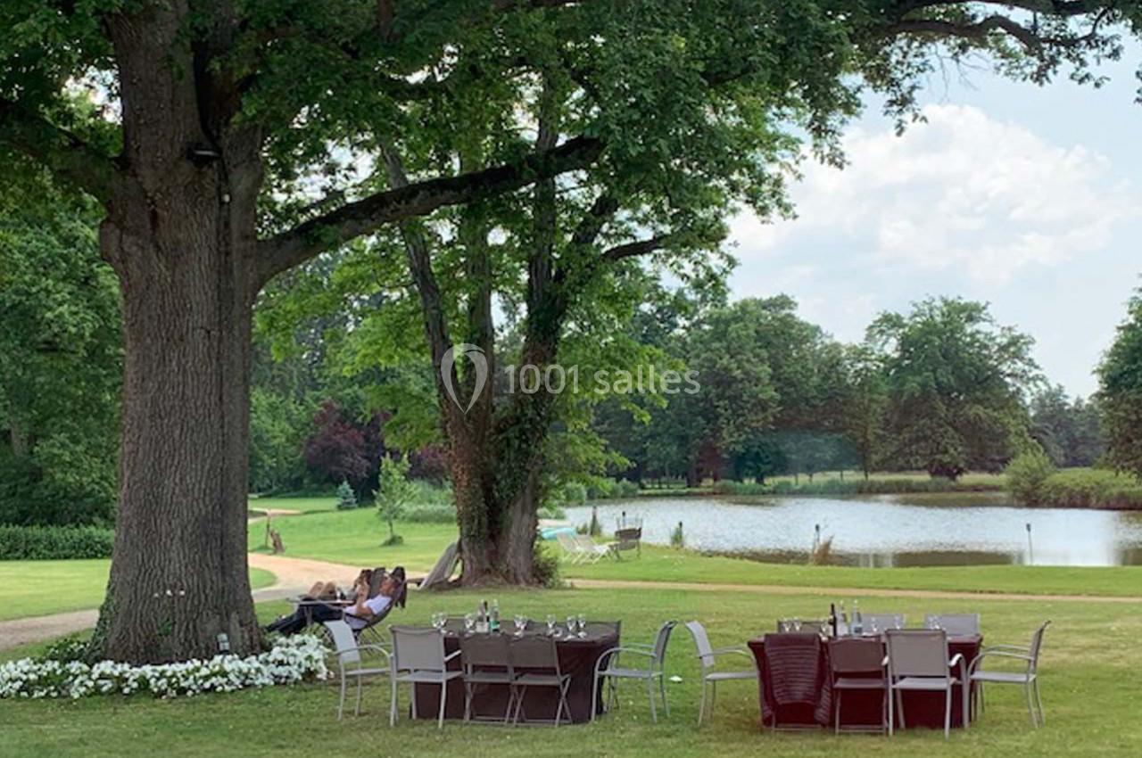 Location salle Cleppé (Loire) - L'Orangerie de Châtel #16 Tables et chaises disposées sur une pelouse ombragée près d'un étang, entourées d'arbres et de verdure.