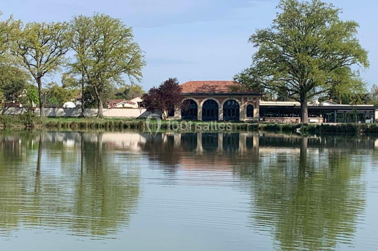 Location salle Cleppé (Loire) - L'Orangerie de Châtel #19 Bâtiment en pierre avec toit en tuiles, bordant un étang entouré d'arbres sous un ciel dégagé.