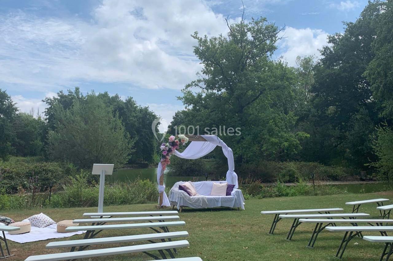 Location salle Cleppé (Loire) - L'Orangerie de Châtel #13 Cérémonie en plein air avec arche fleurie blanche et violette, bancs alignés sur une pelouse près d'un étang.