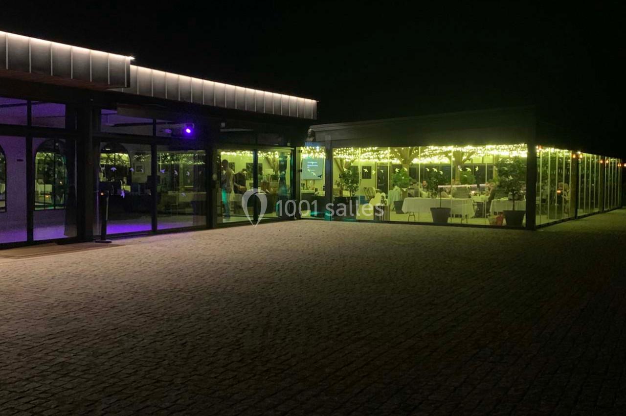 Location salle Cleppé (Loire) - L'Orangerie de Châtel #34 Salle de réception vitrée éclairée de nuit, avec des tables dressées visibles à l'intérieur.