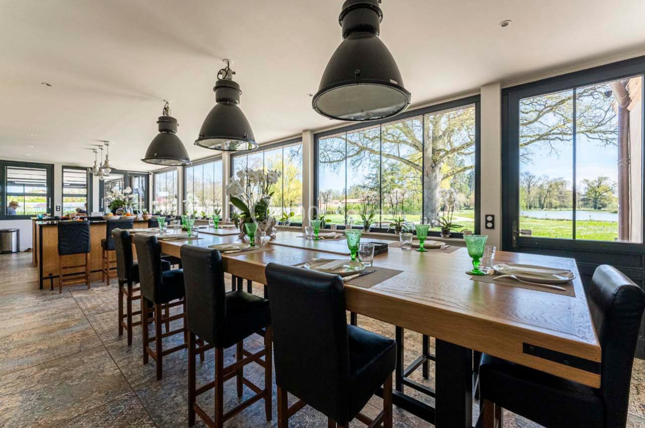 Location salle Cleppé (Loire) - L'Orangerie de Châtel #30 Salle lumineuse avec une grande table en bois, chaises hautes, vaisselle verte et vue sur un paysage verdoyant.