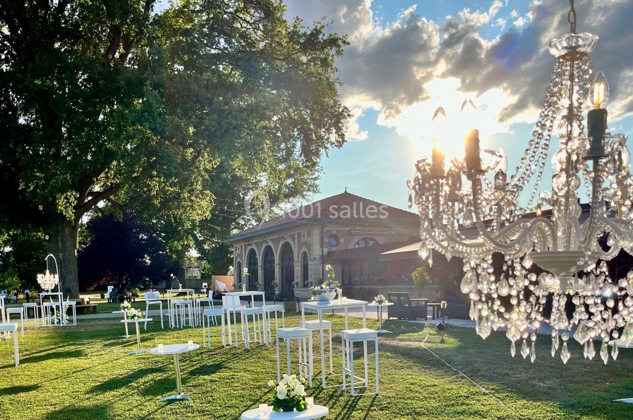 Location salle Cleppé (Loire) - L'Orangerie de Châtel #28 Jardin aménagé pour un événement avec tables hautes, chandeliers et bâtiment en arrière-plan sous un ciel ensoleillé.
