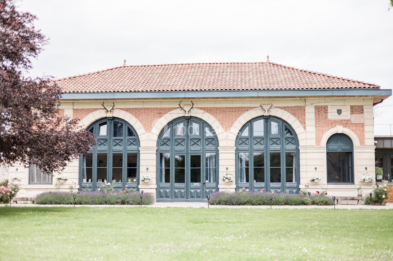 Location salle Cleppé (Loire) - L'Orangerie de Châtel #1 Bâtiment en briques et pierres avec grandes arches vitrées, entouré de pelouse et d'arbres.