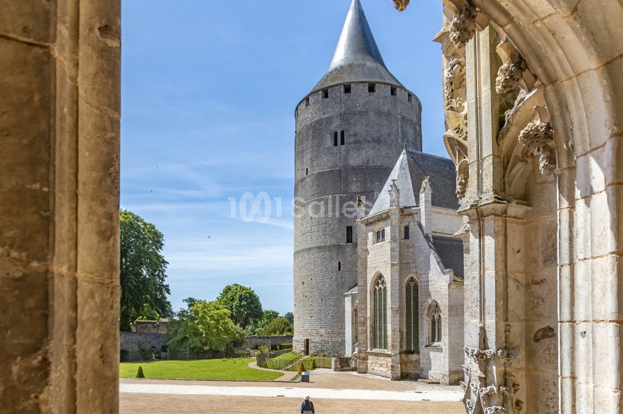 Vue d'une tour médiévale et d'une chapelle entourées de verdure, encadrées par une arche en pierre sculptée.