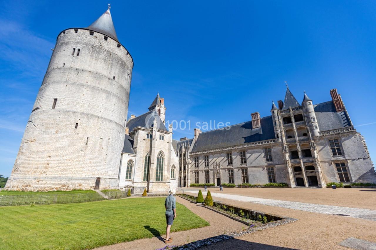 Un homme marche dans la cour d'un château avec une tour ronde et des bâtiments en pierre sous un ciel bleu.
