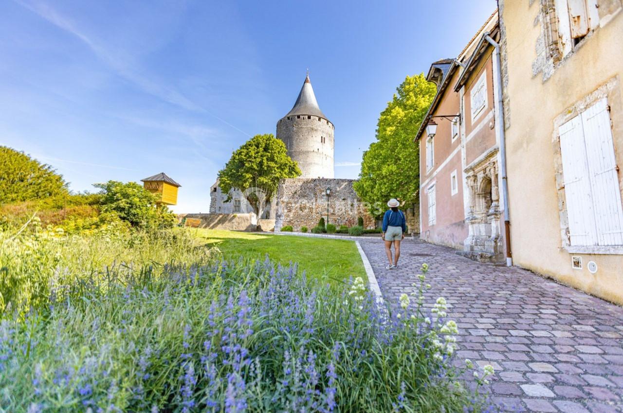 Un homme marche sur une allée pavée bordée de fleurs, menant à une tour médiévale entourée de murs et de maisons anciennes.