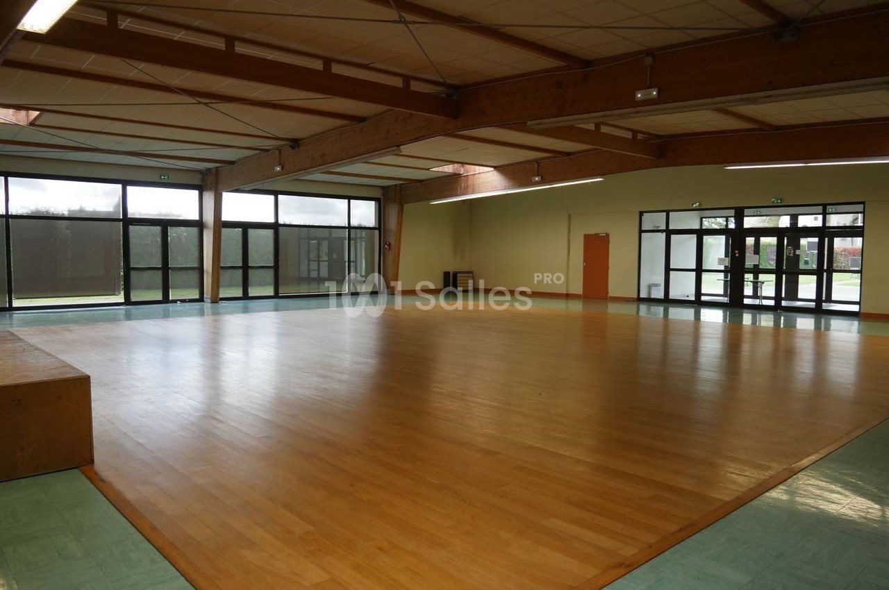 Salle spacieuse avec parquet en bois, murs clairs, grandes baies vitrées et plafond avec poutres apparentes.