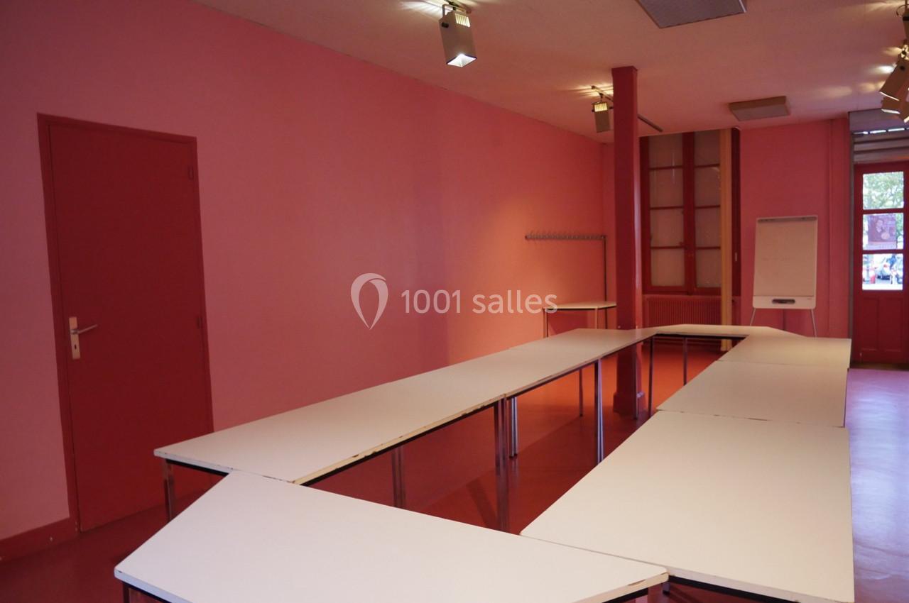 Salle rose avec tables blanches disposées en U, tableau de conférence et porte fermée en arrière-plan.