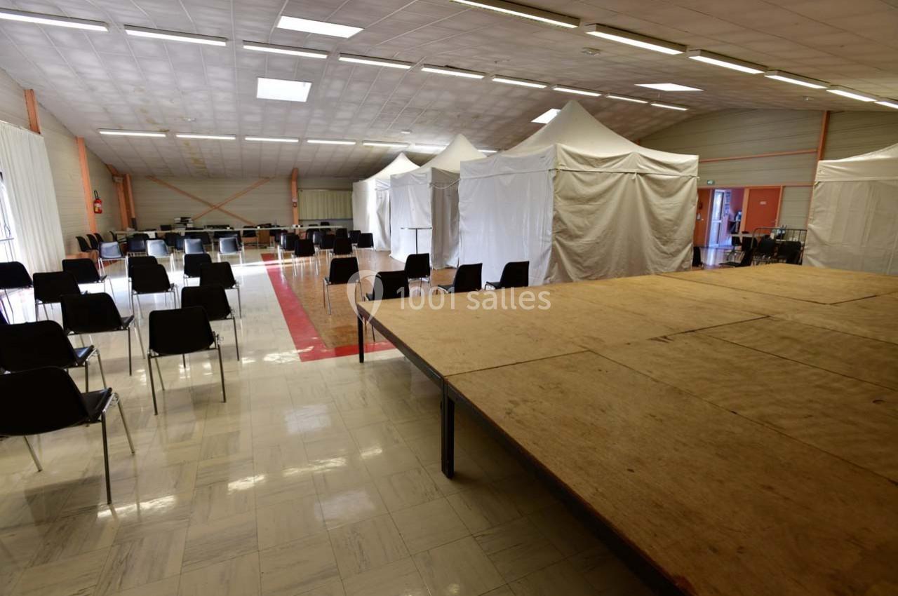 Salle avec des chaises disposées devant une estrade en bois, des tentes blanches alignées en arrière-plan.