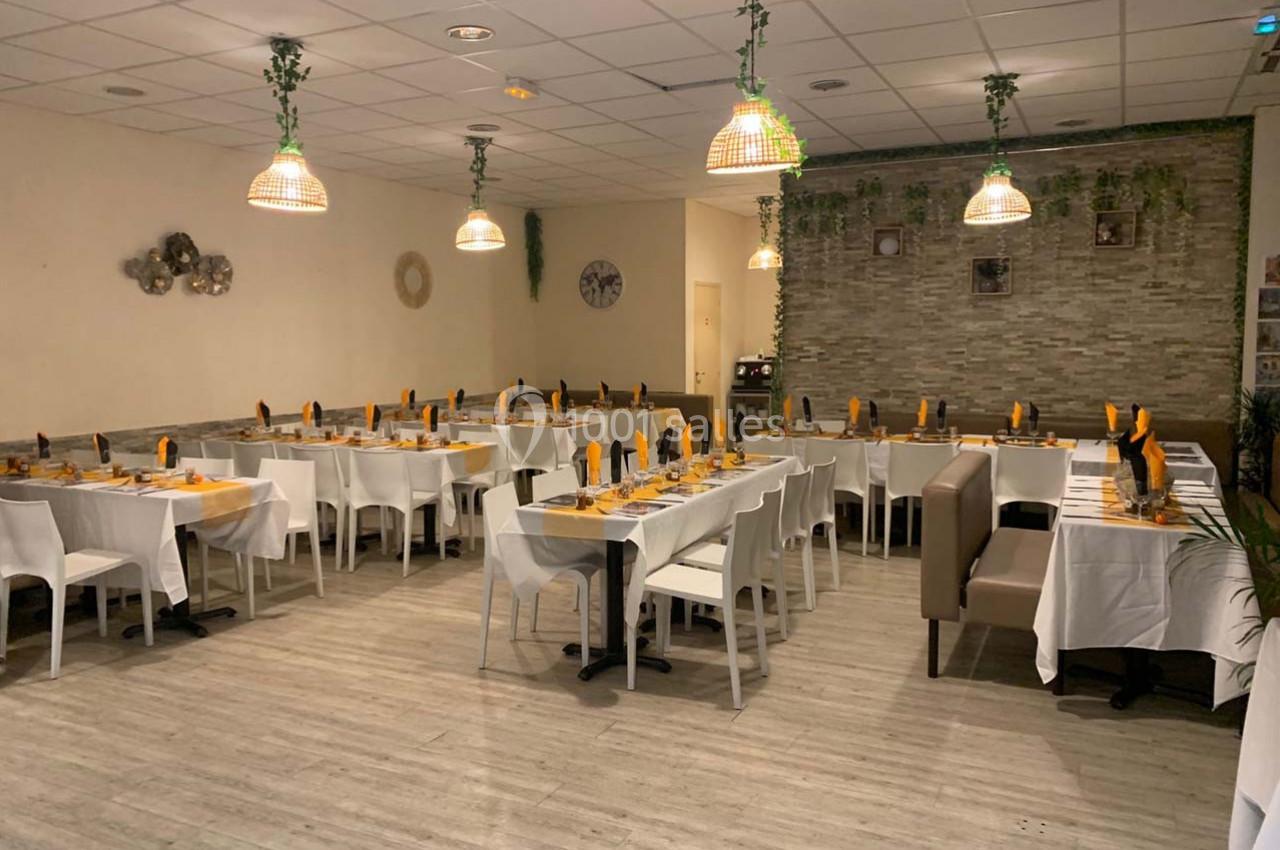 Salle de restaurant décorée avec des tables dressées, nappes blanches, serviettes orange et luminaires suspendus.