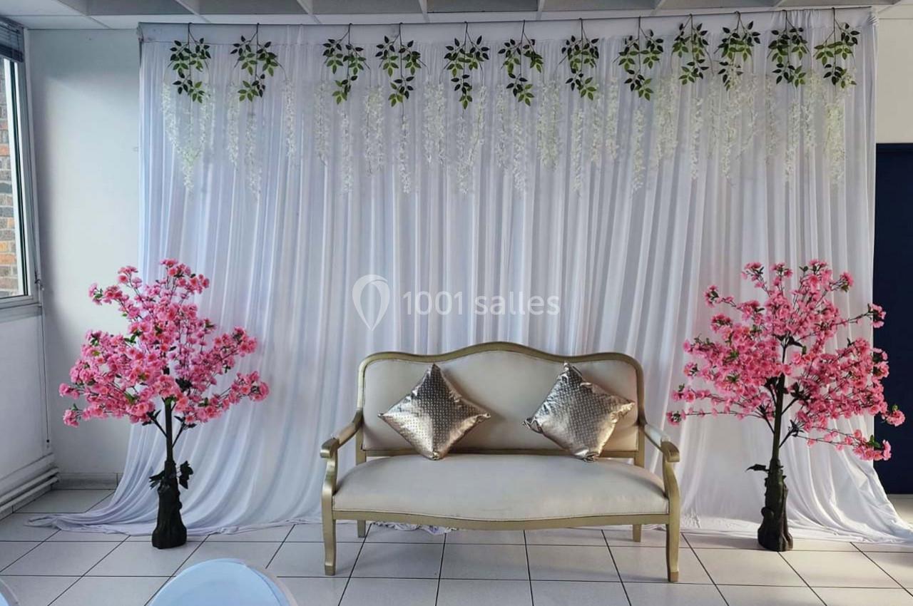 Canapé doré avec coussins argentés, encadré de deux arbres roses, devant un rideau blanc décoré de feuillages.