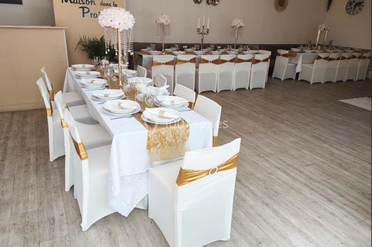Salle de réception décorée avec des tables dressées, nappes blanches, chemins de table dorés et chaises ornées de rubans…