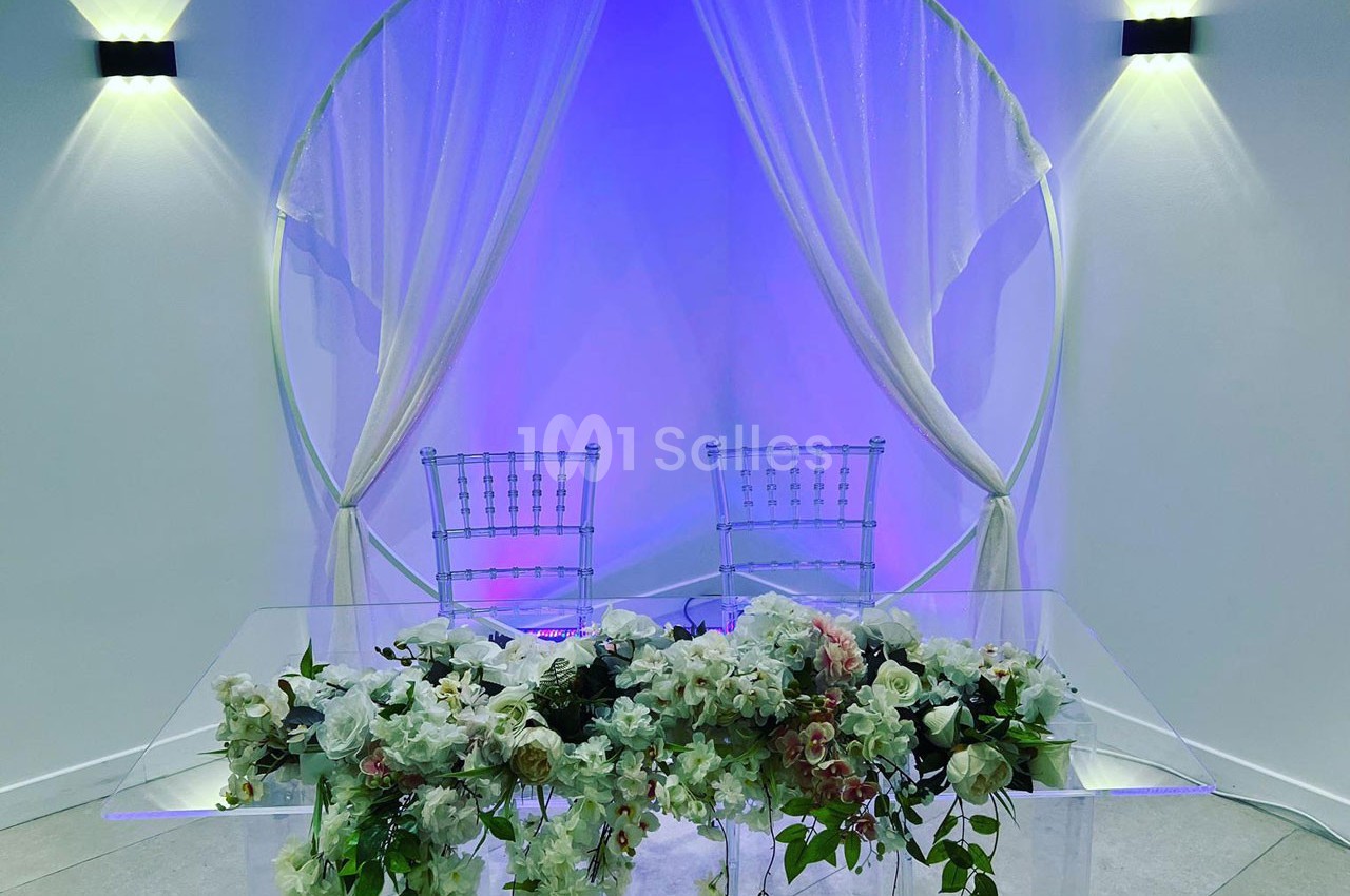 Deux chaises transparentes derrière une table ornée de fleurs blanches et roses, devant un fond lumineux bleu.