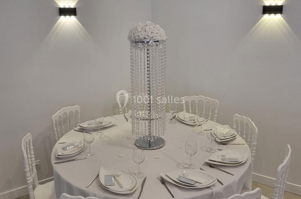 Table ronde décorée pour un événement, avec nappes blanches, vaisselle élégante et centre de table orné de perles et fleurs.