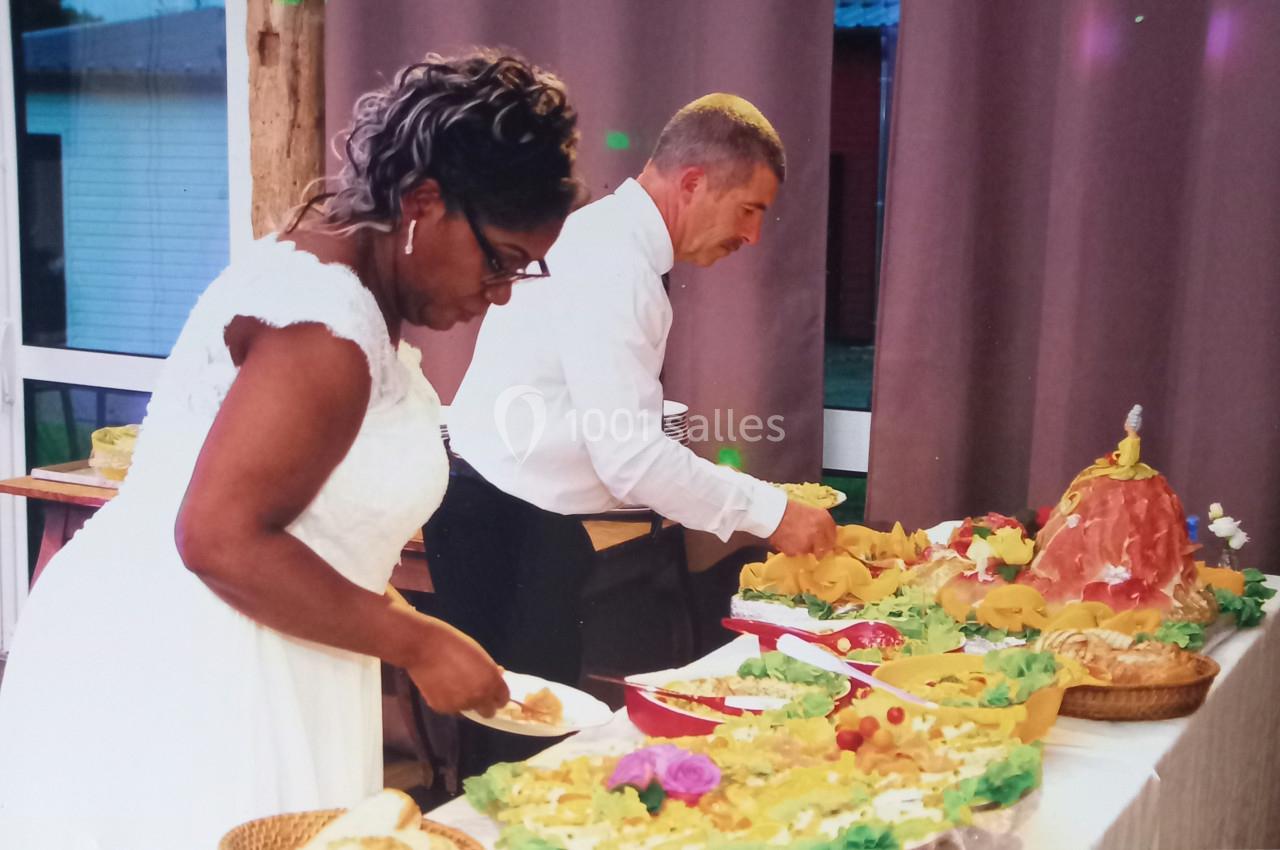 Un couple sert des plats variés sur un buffet décoré, dans une salle avec rideaux violets en arrière-plan.