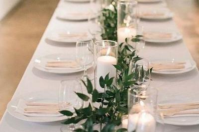 Table élégamment dressée avec nappes blanches, chemins de table verts, serviettes pliées et décorations florales.