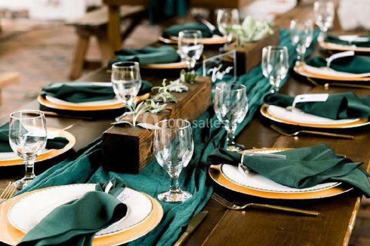 Table en bois dressée avec des assiettes blanches, serviettes vertes, verres et décoration végétale centrale.
