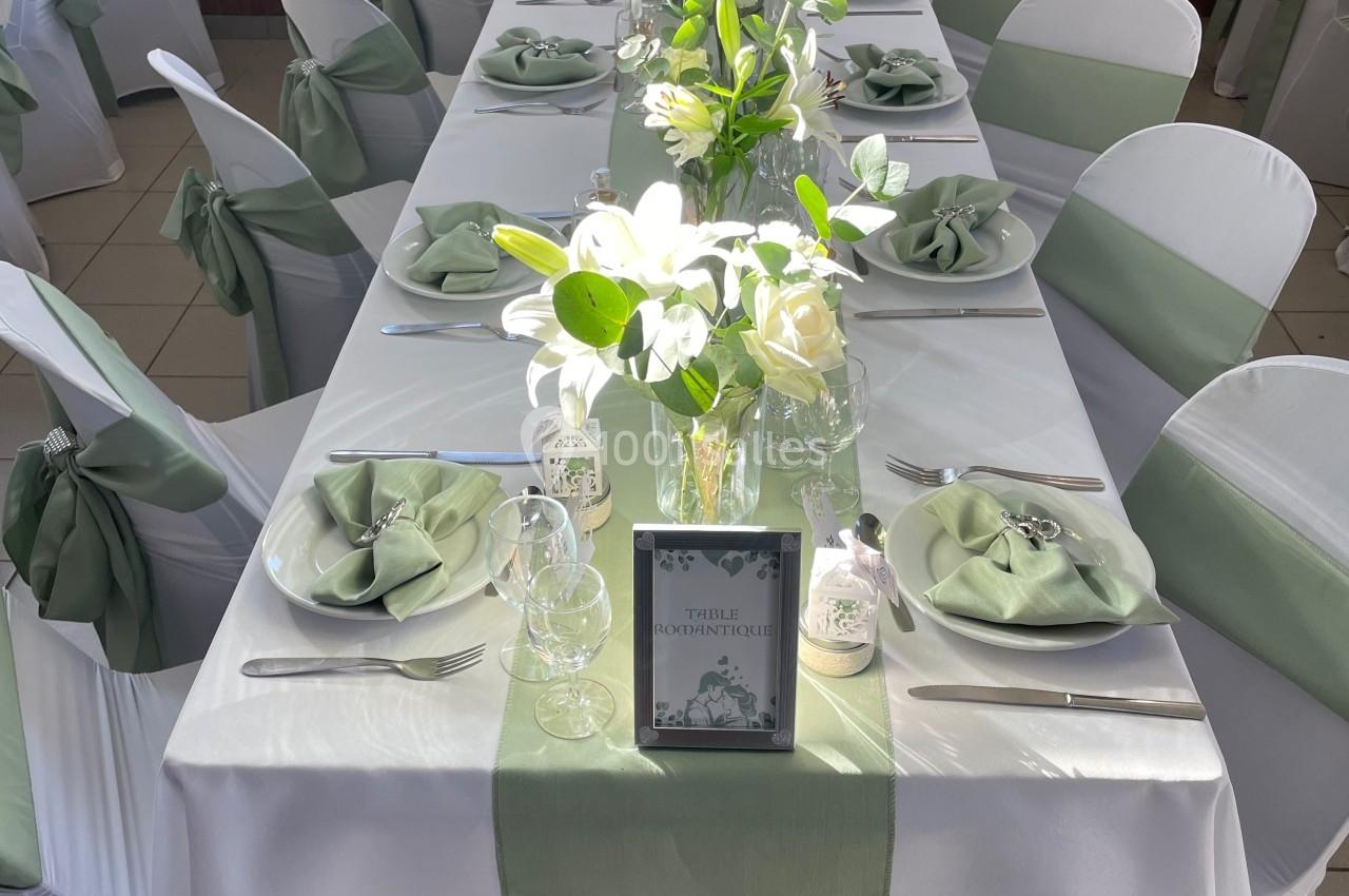 Table décorée avec nappes blanches, chemins de table verts, serviettes pliées, verres et arrangements floraux.