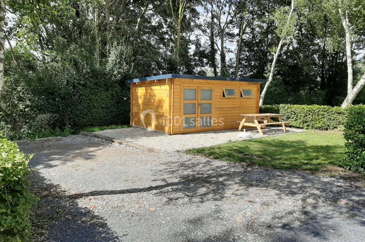 Abri en bois avec fenêtres, entouré de végétation, situé sur un terrain gravillonné avec une table de pique-nique.