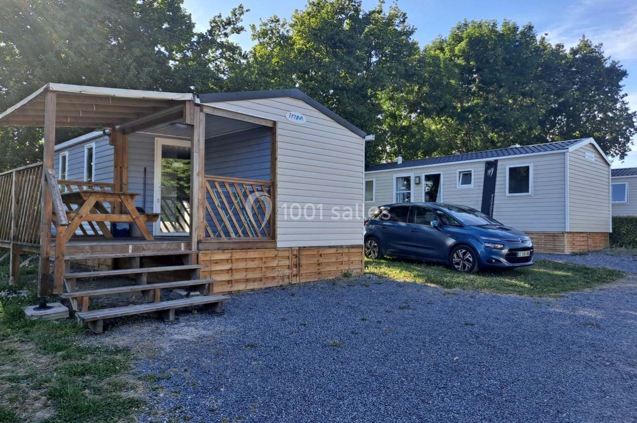 Deux mobil-homes avec terrasse en bois sur un terrain gravillonné, entourés d'arbres et d'espaces verts.