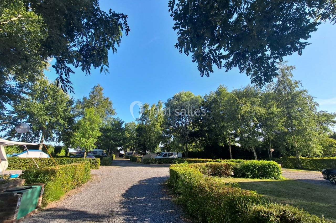 Allée gravillonnée bordée de haies et d'arbres, menant à des emplacements de camping sous un ciel dégagé.