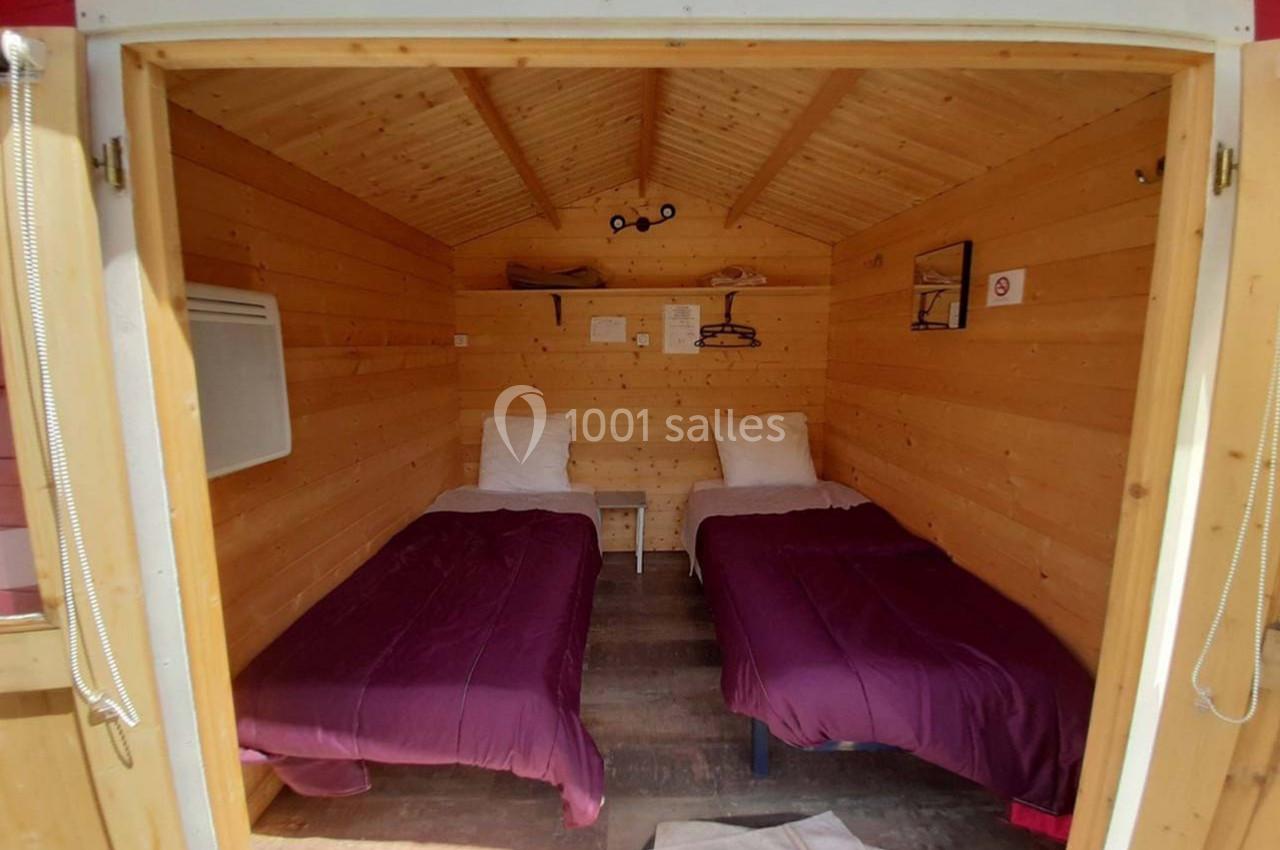 Deux lits simples avec draps violets dans une petite cabane en bois, éclairée par la lumière naturelle.