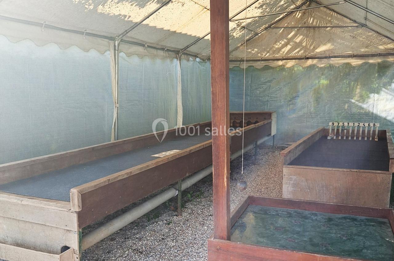 Piste de jeu de quilles en bois sous une tente, avec des quilles alignées et un sol en gravier.