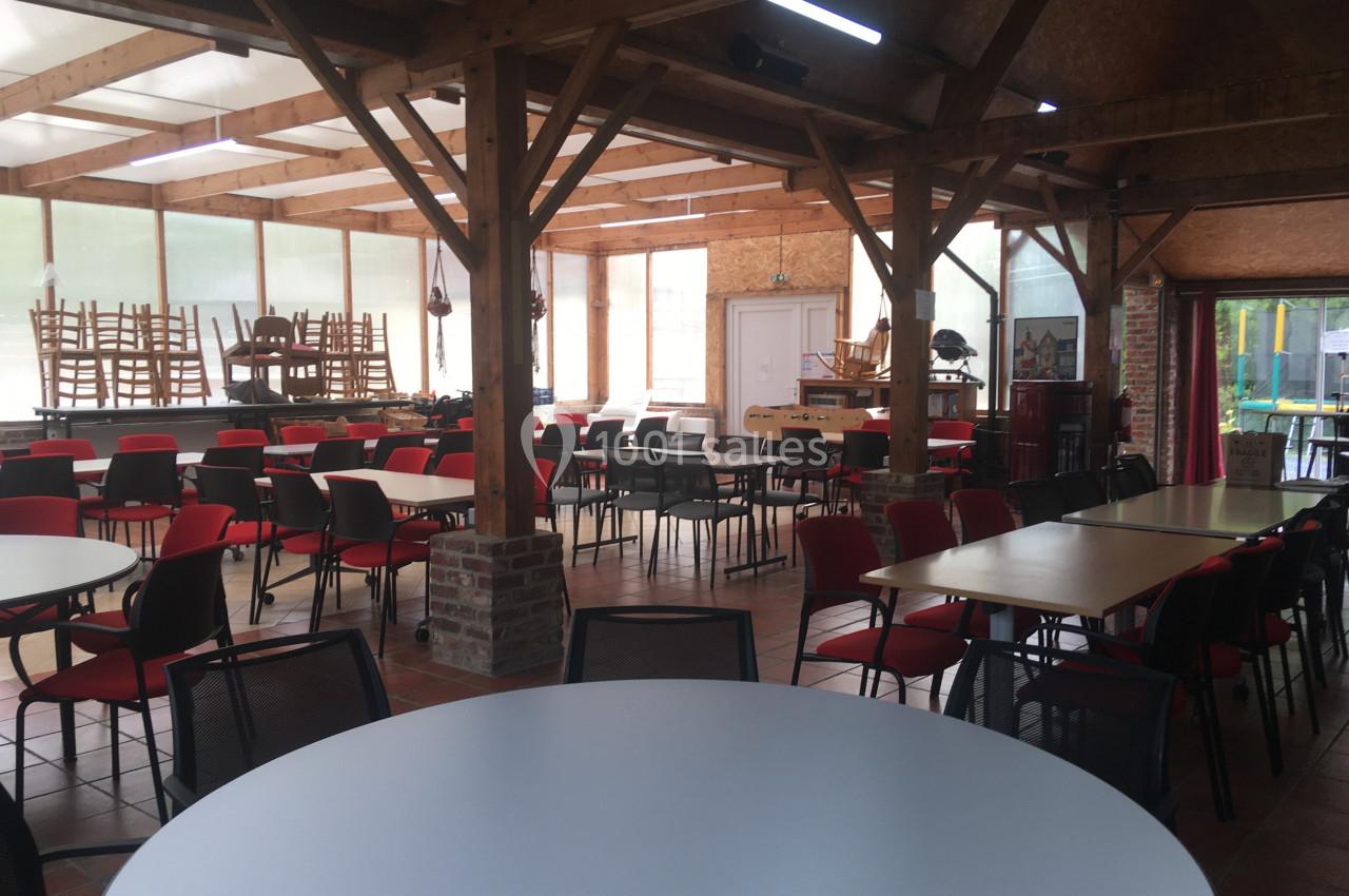 Salle lumineuse avec des tables rondes et rectangulaires, chaises empilées et structure en bois apparent.