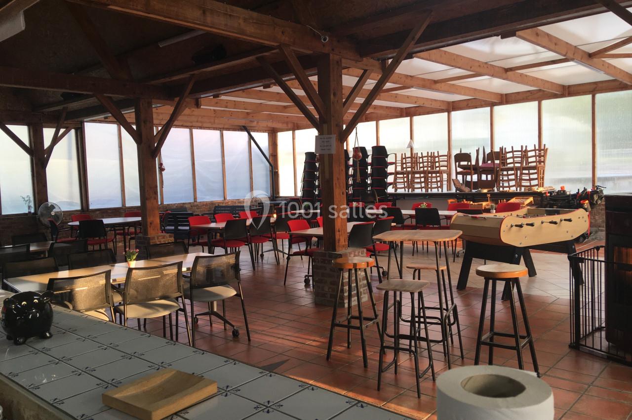Salle en bois avec tables et chaises disposées, certaines empilées, éclairée par la lumière naturelle à travers de grandes…