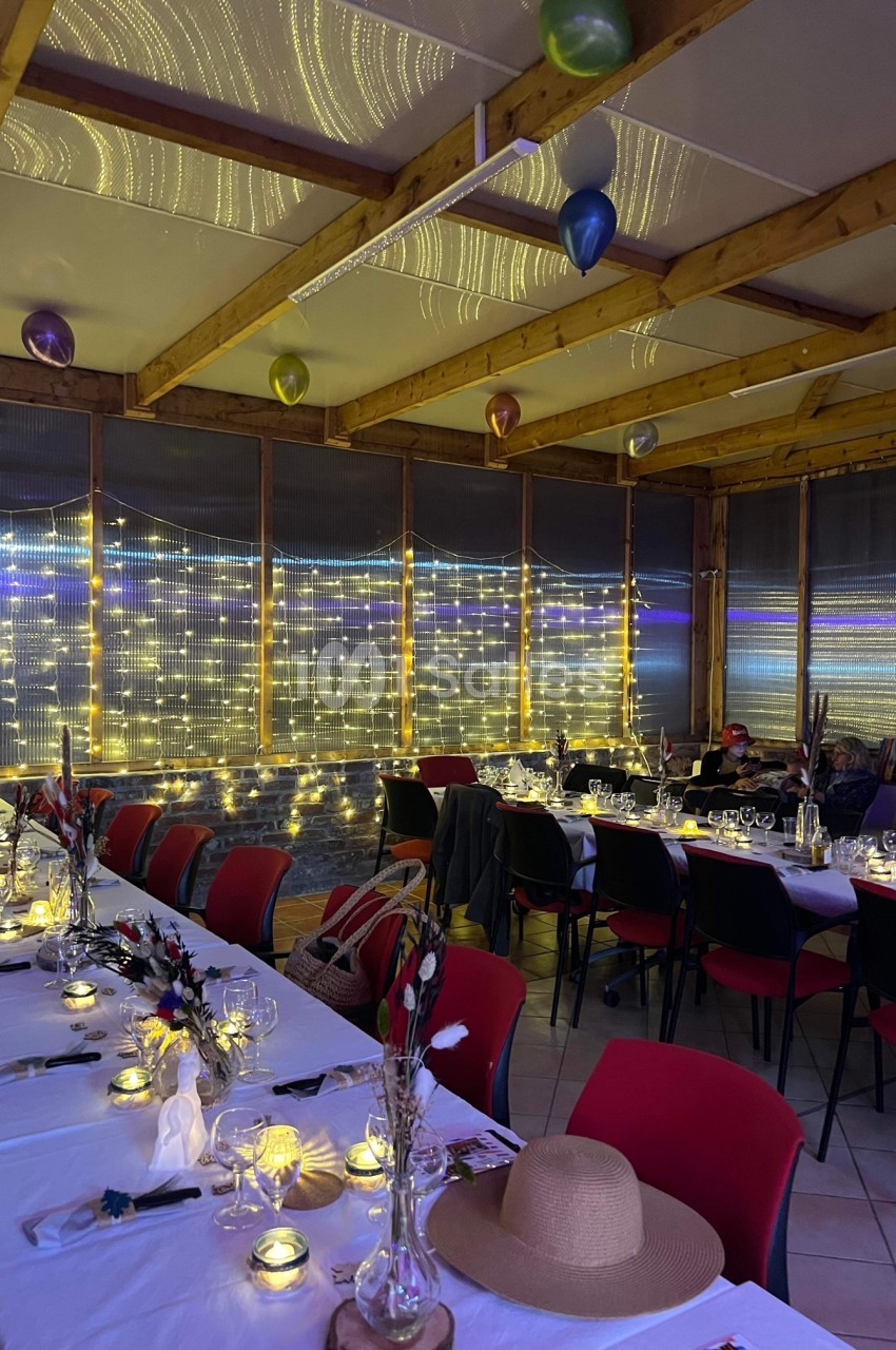 Salle décorée pour un événement, avec tables dressées, guirlandes lumineuses et ballons suspendus au plafond.