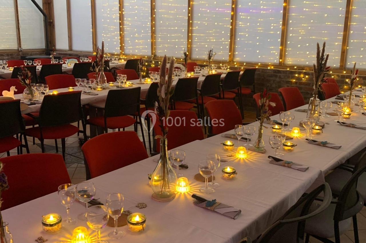 Salle décorée pour un repas, avec longues tables dressées, bougies allumées et guirlandes lumineuses en arrière-plan.