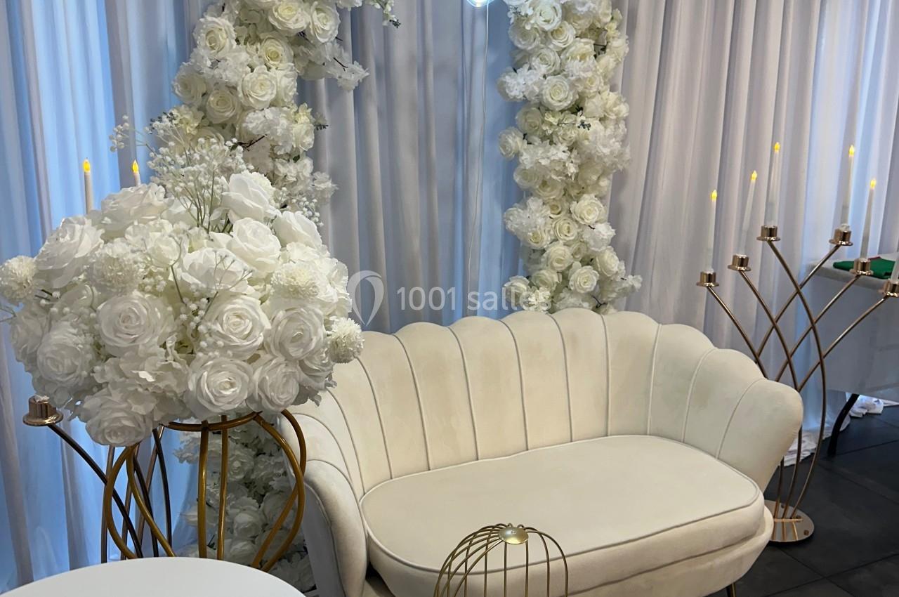 Canapé beige devant un rideau blanc, décoré d'arcs de fleurs blanches, bougies et initiales lumineuses en arrière-plan.