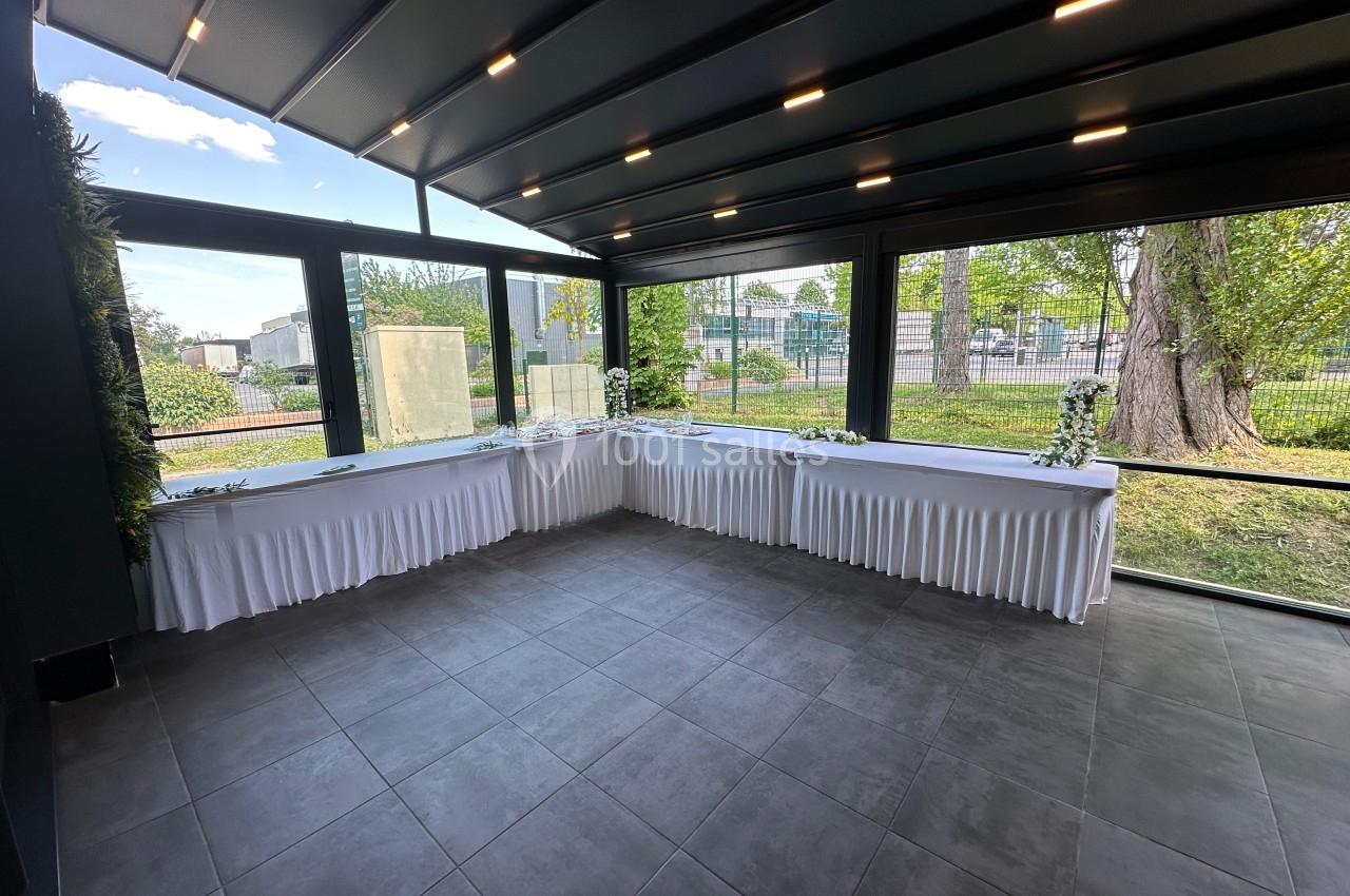 Salle lumineuse avec grandes baies vitrées, tables drapées de blanc en angle, vue sur un jardin extérieur.