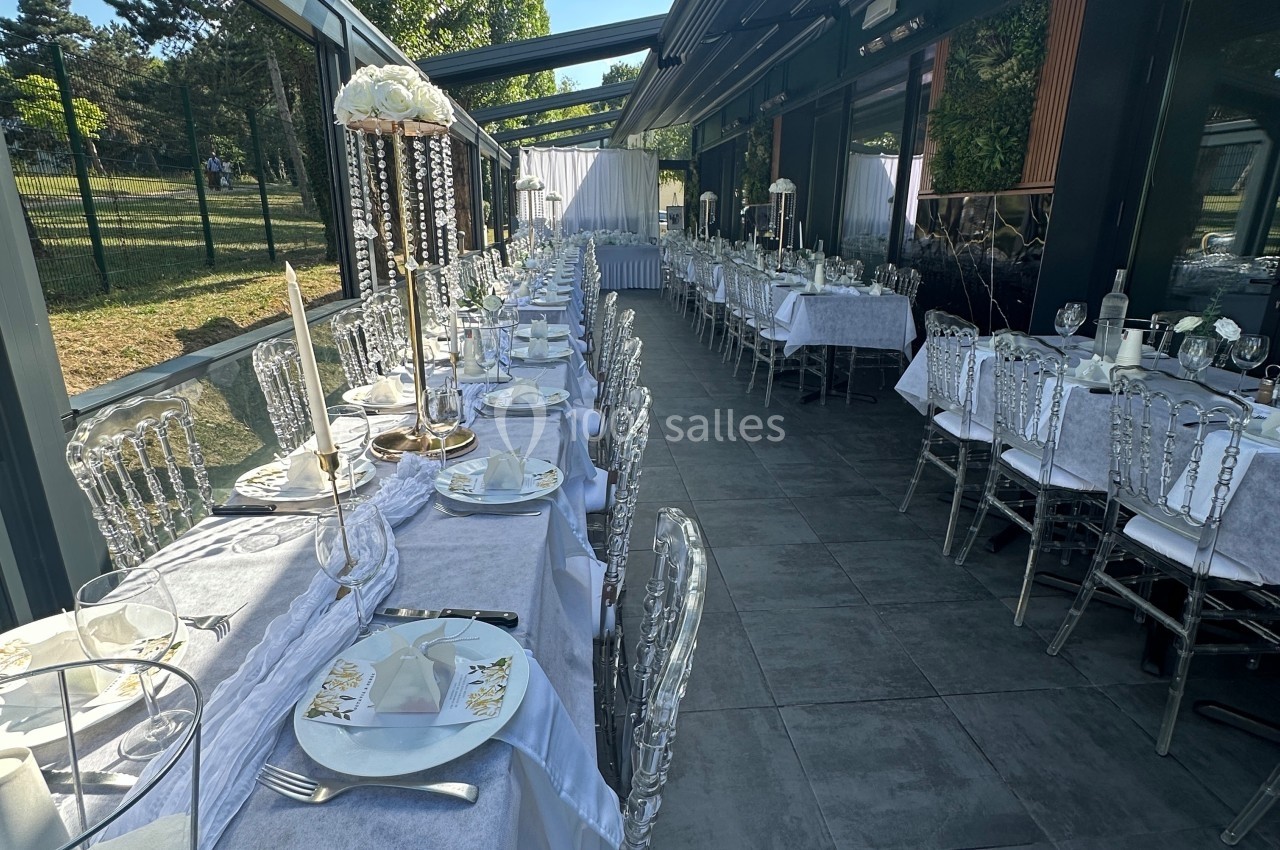 Salle de réception extérieure avec tables dressées, nappes blanches et chaises transparentes sous une verrière.
