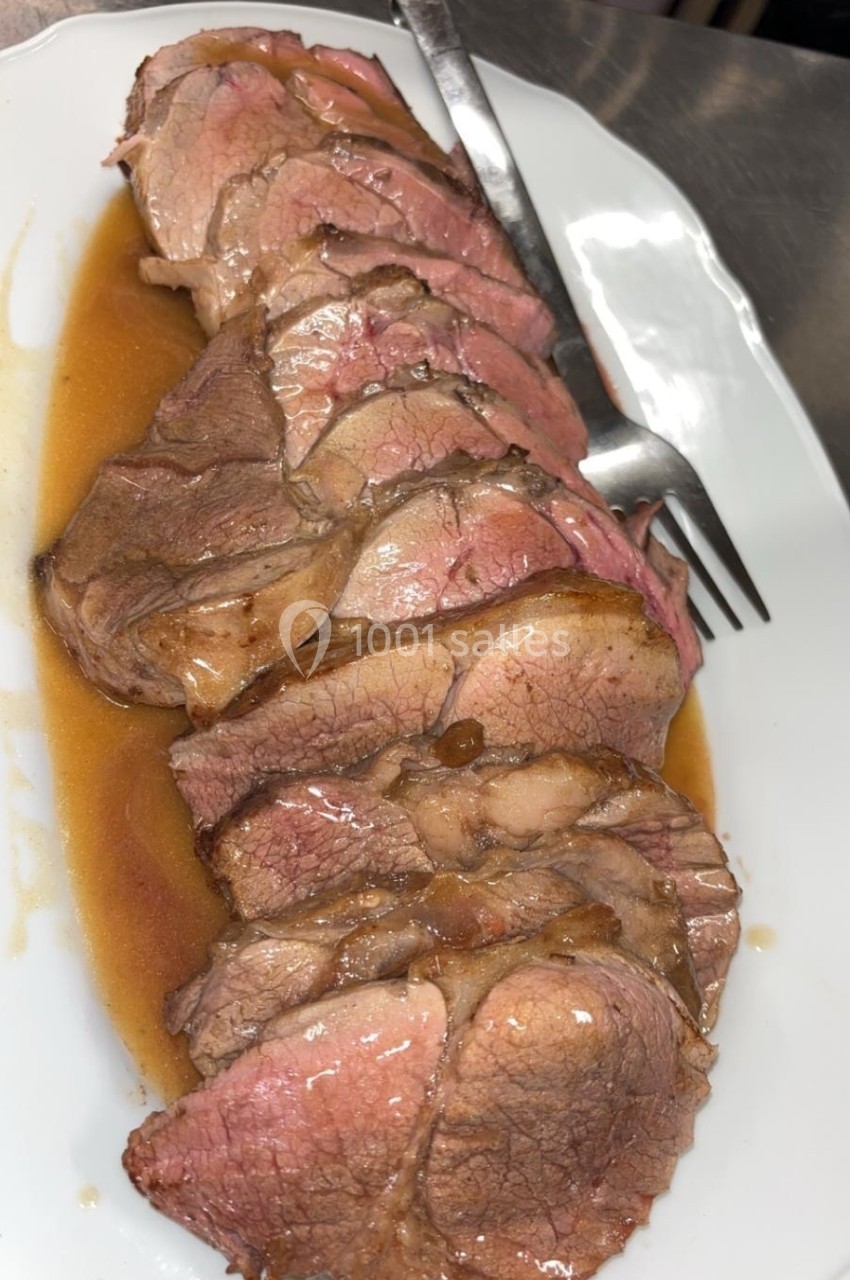 Tranches de viande rôtie servies sur un plat blanc avec une sauce et une fourchette en arrière-plan.