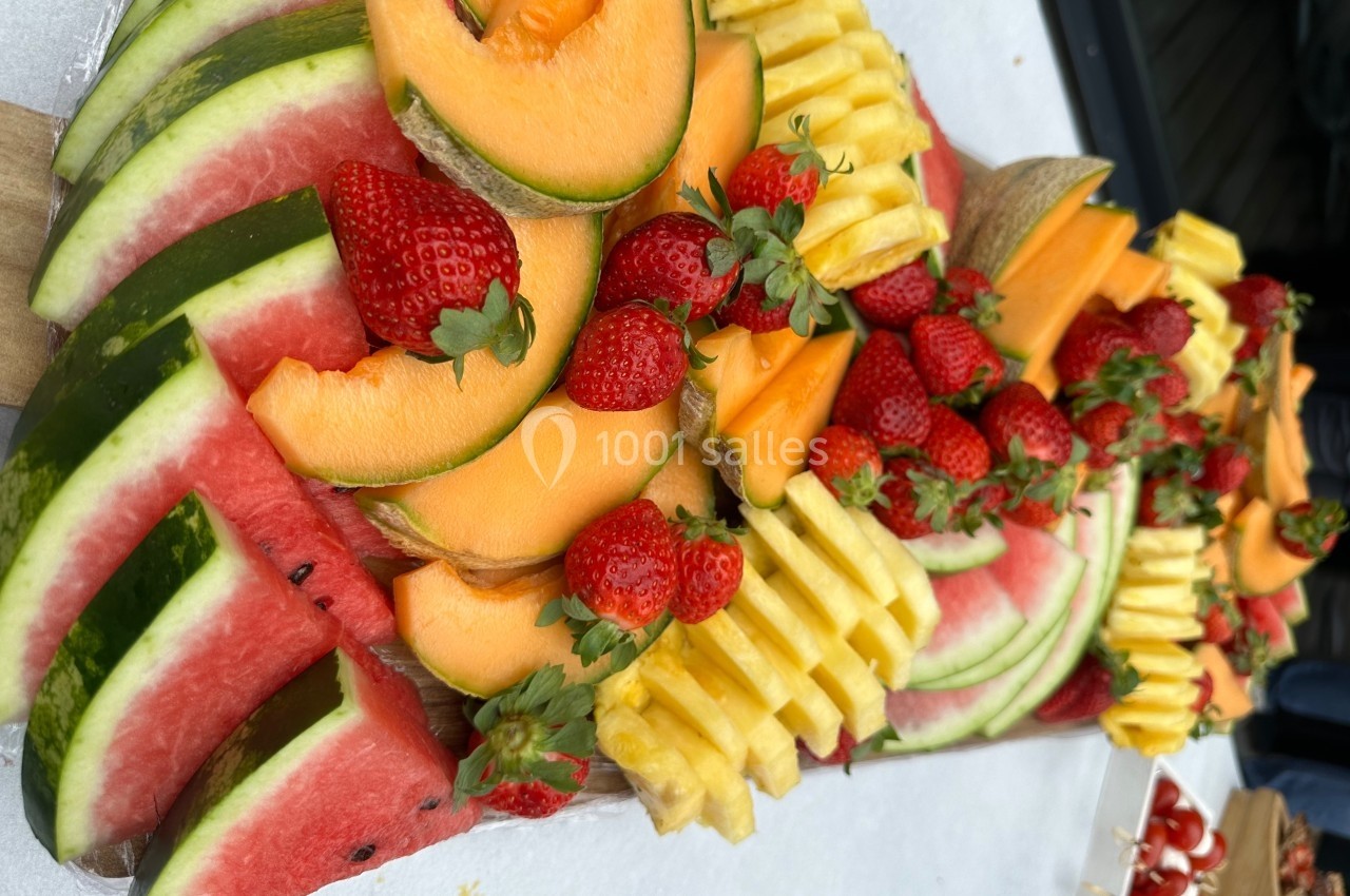 Plateau de fruits frais comprenant pastèque, melon, ananas et fraises, disposés sur une table blanche.