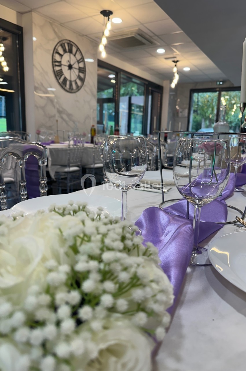 Table élégamment dressée avec des verres, un bouquet de fleurs blanches et une nappe violette dans une salle lumineuse.